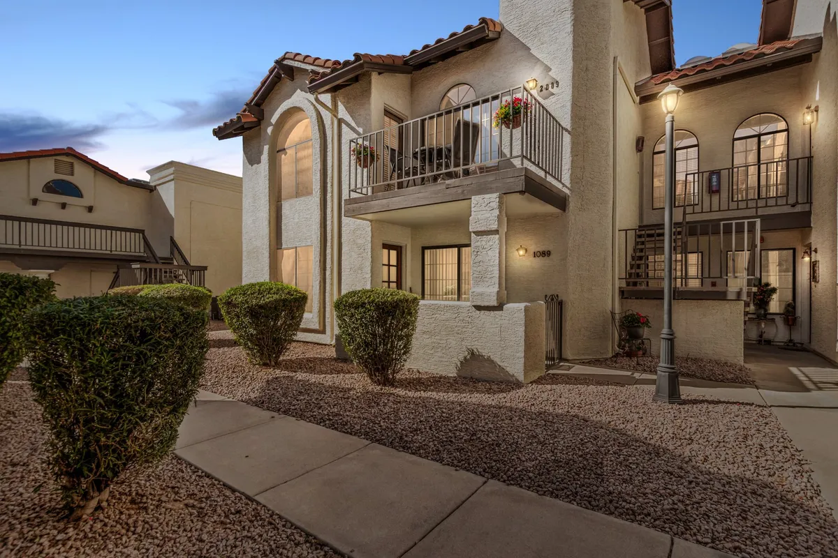 The image shows the elegant exterior of a Scottsdale apartment building in the La Contessa community, with vibrant flowers adorning a private balcony, lush hedges framing the entrance, and appealing architectural details that reflect a warm, inviting atmosphere.