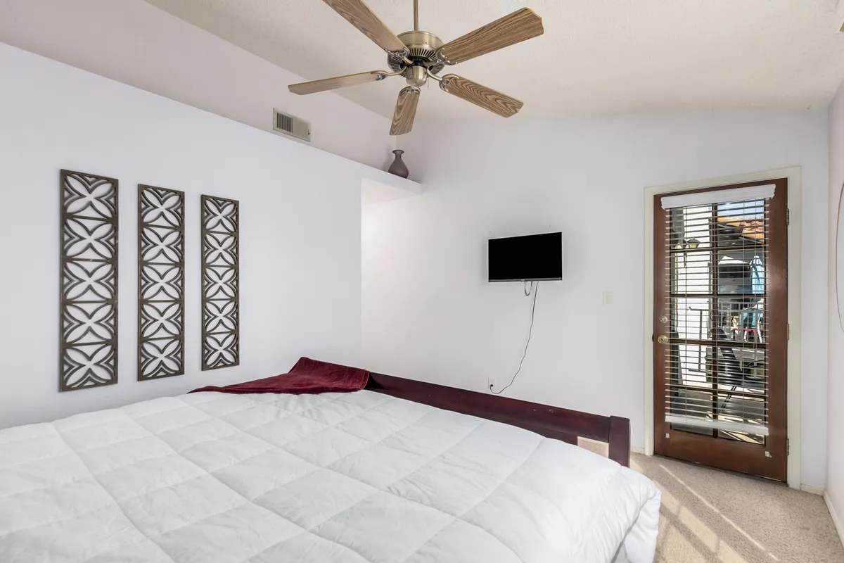 A serene bedroom featuring white bedding, decorative wall panels, a wall-mounted TV, and a door leading directly outside. Natural light pours in from the door, hinting at a private balcony or outdoor space just beyond.