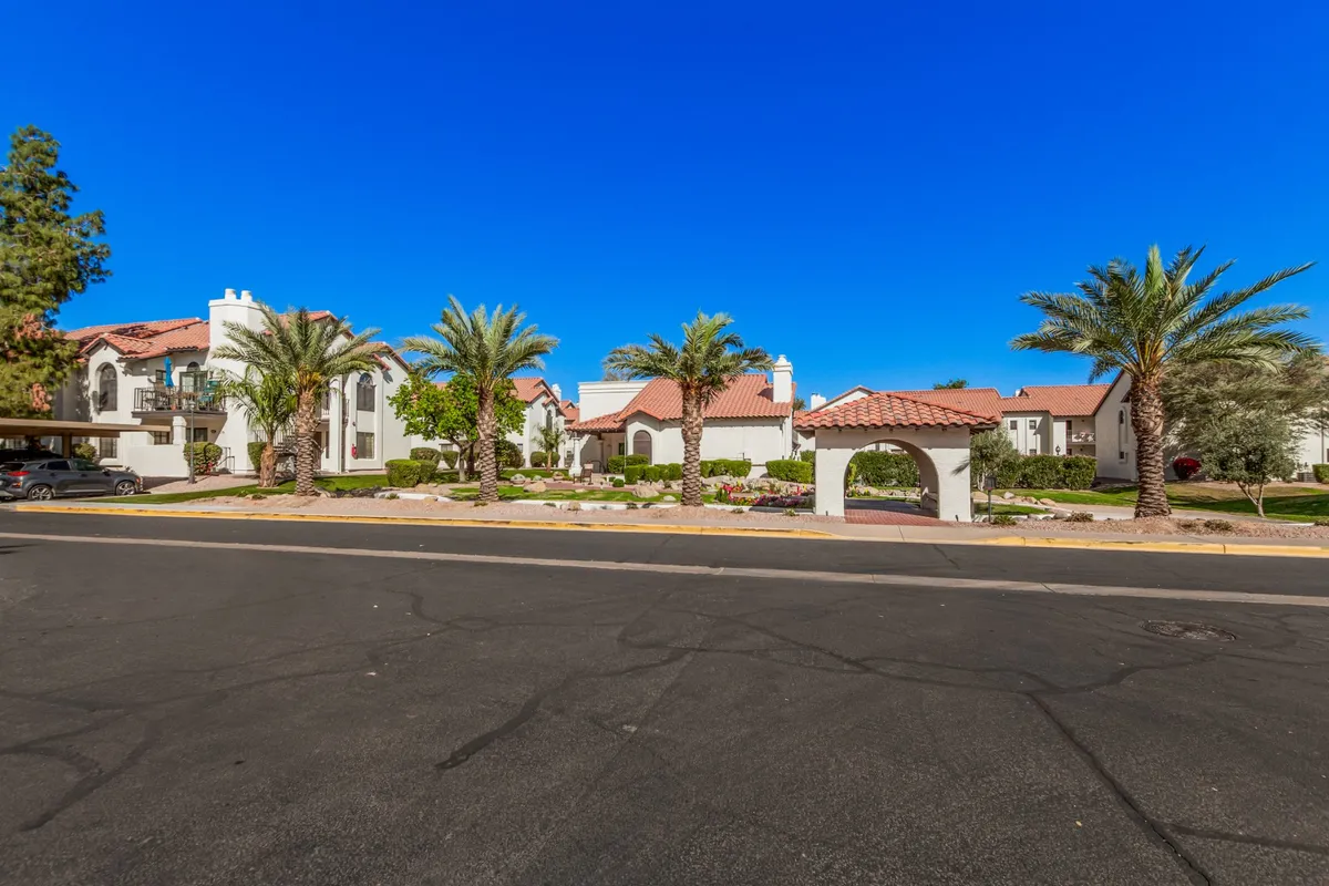 This image captures an inviting exterior scene with a palm-lined entrance, distinctive red tiled roofs, and manicured gardens welcoming residents and visitors. The archway suggests a secure, gated environment, while the lush landscaping provides a sense of community and relaxation.