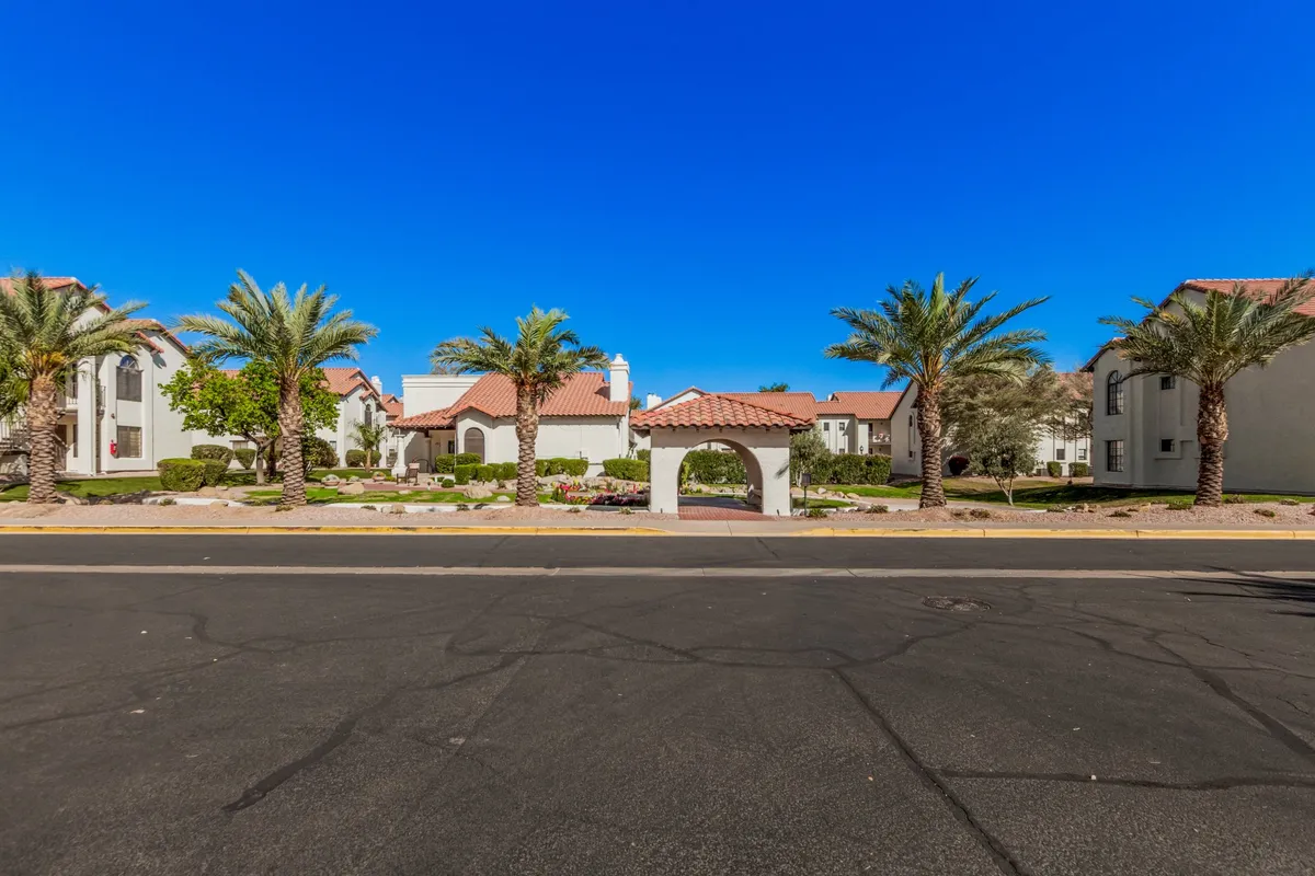 This exterior image highlights the lush landscaped gardens, towering palm trees, and the arched entrance to the community. The white and red buildings in the backdrop evoke the character of La Contessa, while the gardens suggest shared spaces for socializing and relaxation.