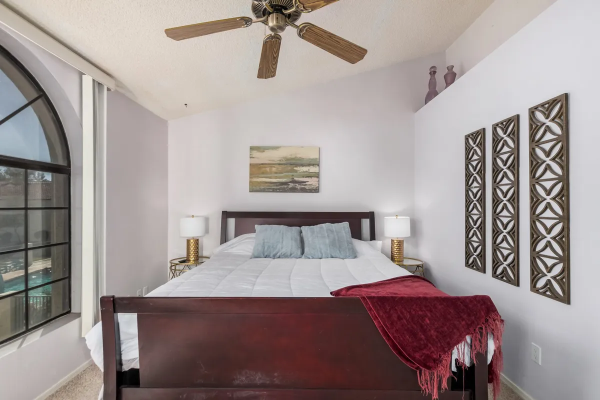 This image presents a spacious bedroom with a plush bed, decorative wall art, large windows letting in natural light, nightstands, and a ceiling fan. The room exudes comfort and serenity, emphasizing a restful atmosphere.