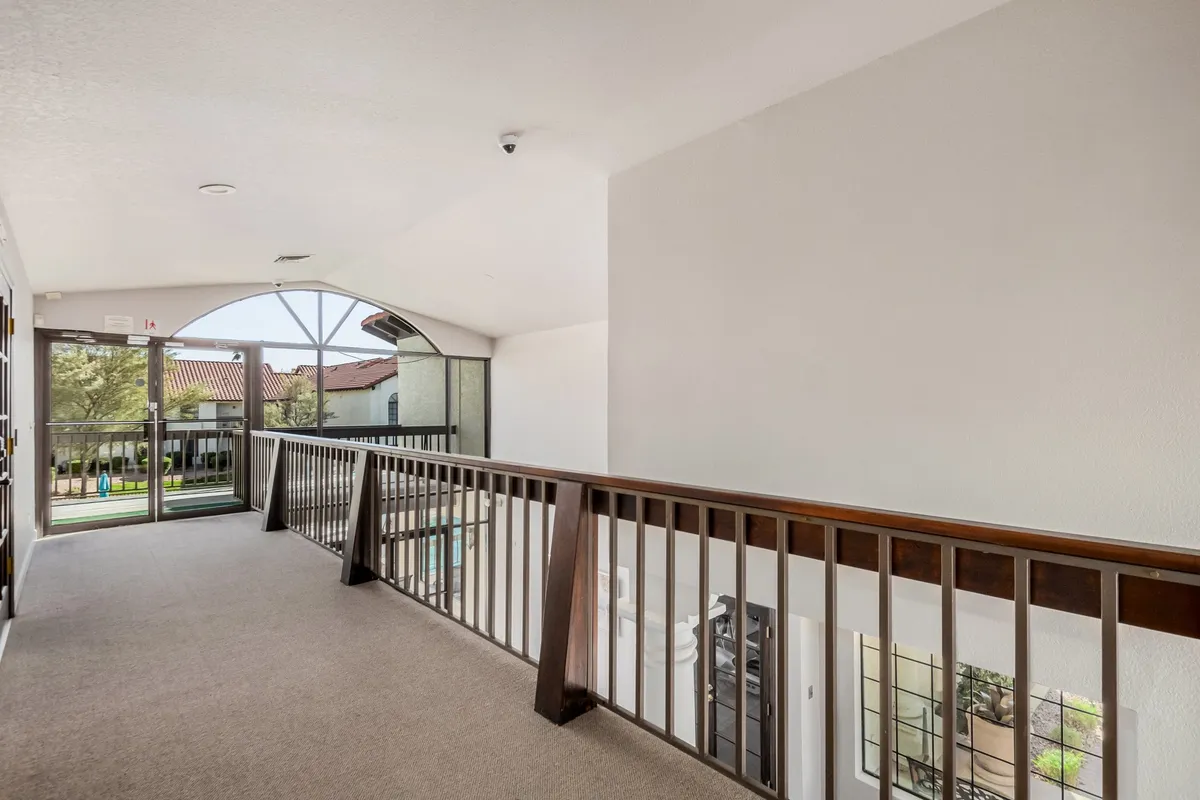 A tranquil hallway with a wooden railing, an arched window letting in natural light, and an inviting entrance door. The scene conveys a sense of peacefulness and spaciousness, with the view from the window suggesting a connection to the outside community.