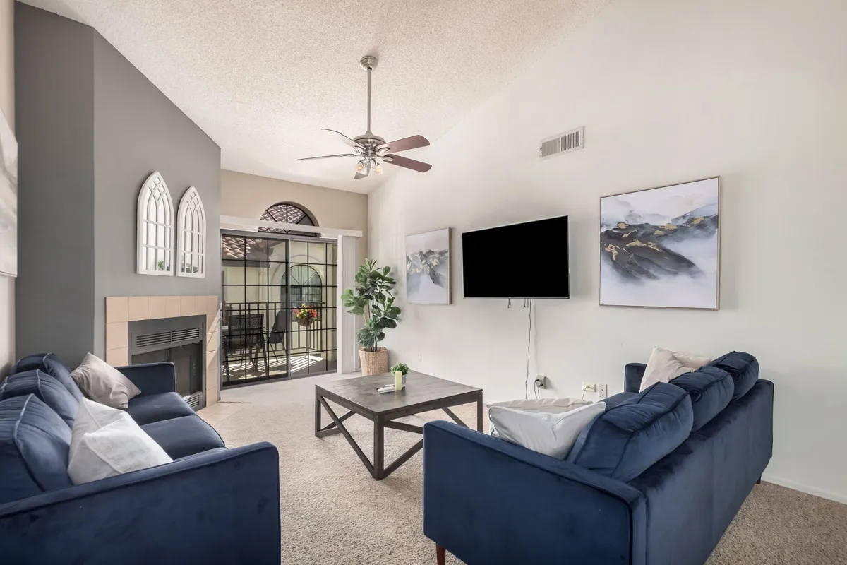 The image showcases a spacious living room featuring blue sofas arranged around a coffee table, a cozy fireplace, large windows letting in abundant sunlight, and decorative plants that soften the space. The arrangement is inviting and perfect for relaxation or social gatherings.