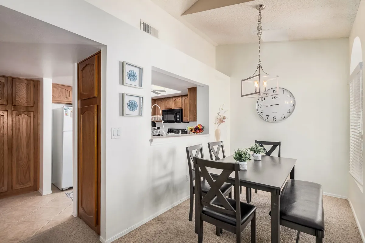 An inviting dining area with a large table surrounded by chairs, a clock on the wall, a kitchen pass-through that connects to the kitchen, and decorative art accents. The space is set up for family meals, casual gatherings, or even homework, with the open design fostering conversation and connection.