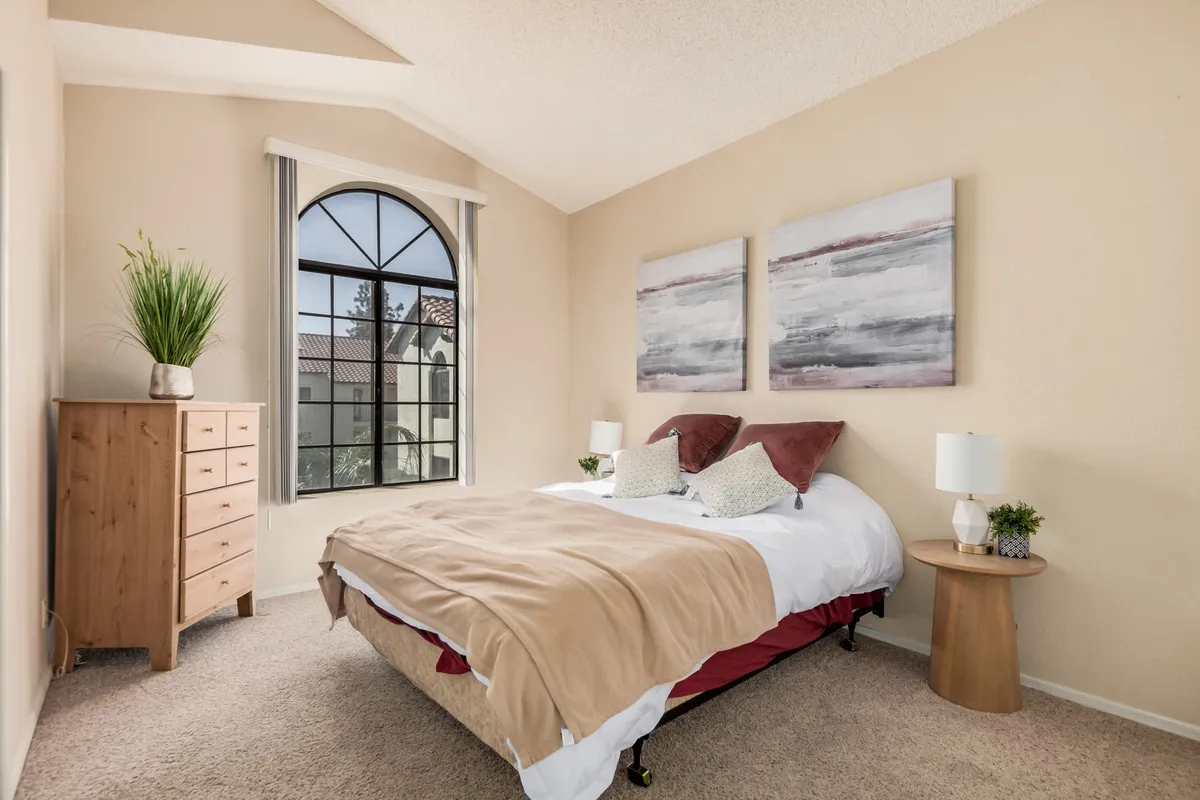 The photo features a bed with neutral bedding, tasteful artwork above, a stylish arched window, and a nightstand with a lamp. The bedroom is cozy and well-lit, with an intimate, calming mood.
